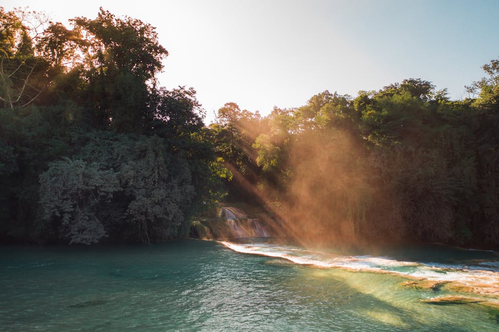 A peaceful waterfall cascades amidst lush forest with beams of sunlight streaming through, creating a tranquil scene.