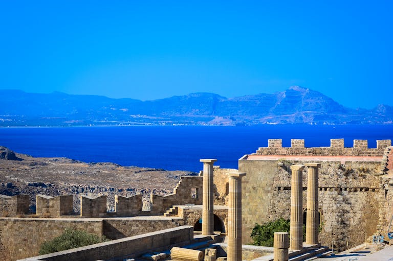 Scenic view of ancient Greek ruins with stone columns and walls against a vibrant blue Aegean Sea backdrop.