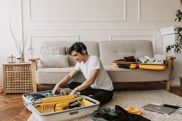 Young man in a living room, packing a suitcase while wearing headphones and listening to music.