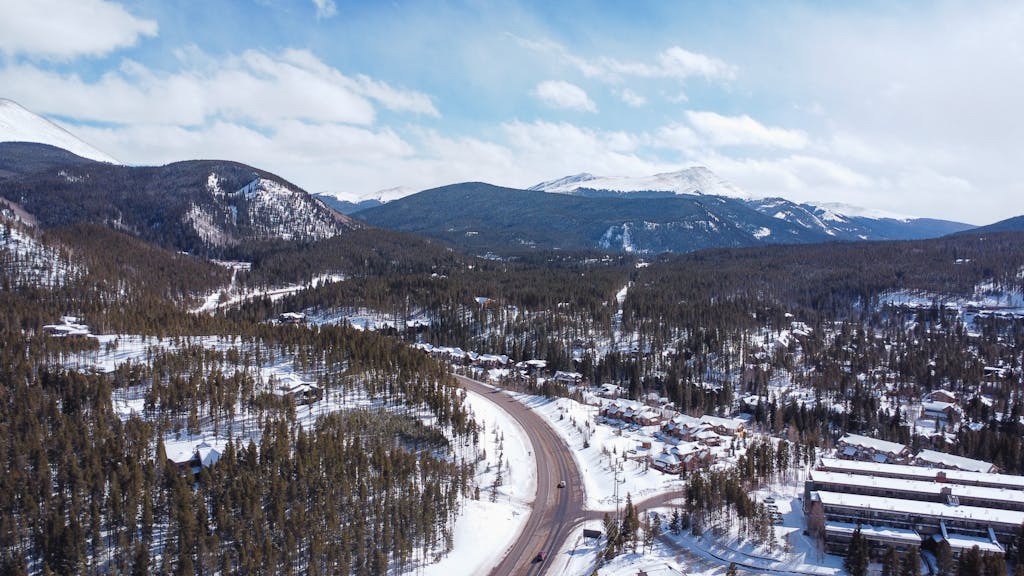 A breathtaking aerial view of a snow-covered mountain road winding through evergreen forests in winter.