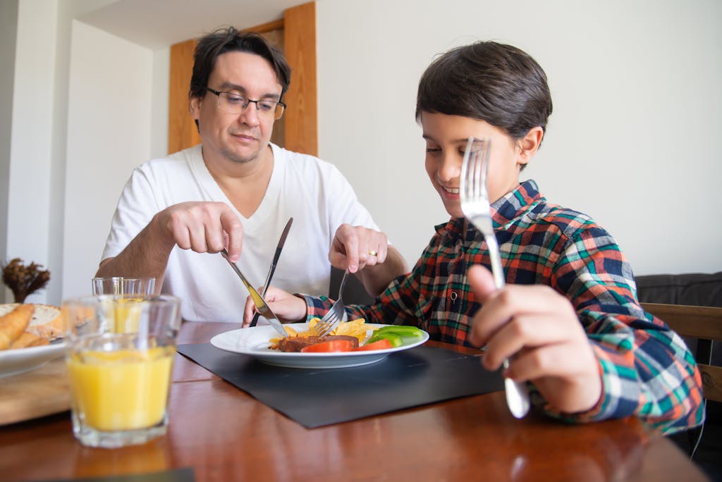 A father and son share a meal indoors, practicing dining etiquette with orange juice and healthy options.