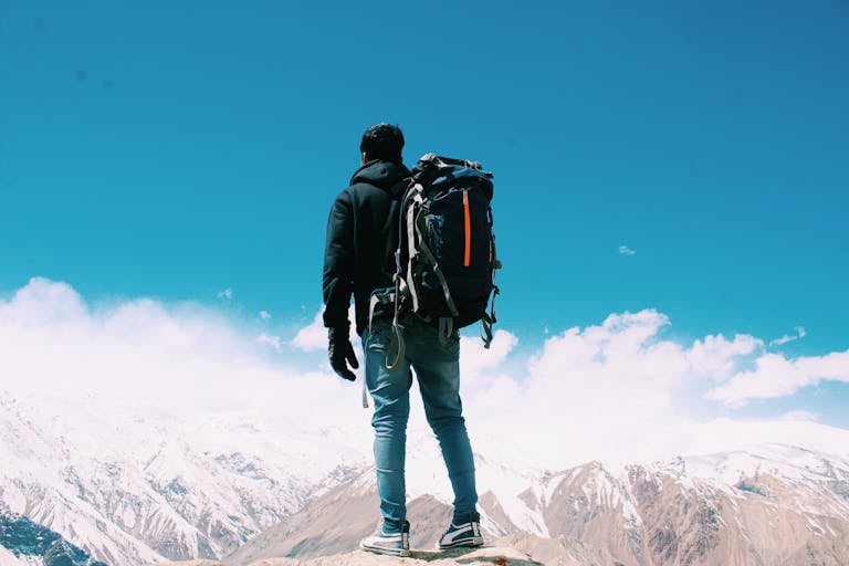 A lone hiker with a backpack surveys snow-capped mountains under a clear blue sky.