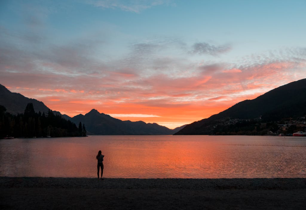 A person stands silhouetted against a stunning sunset over a tranquil lake with distant mountains.