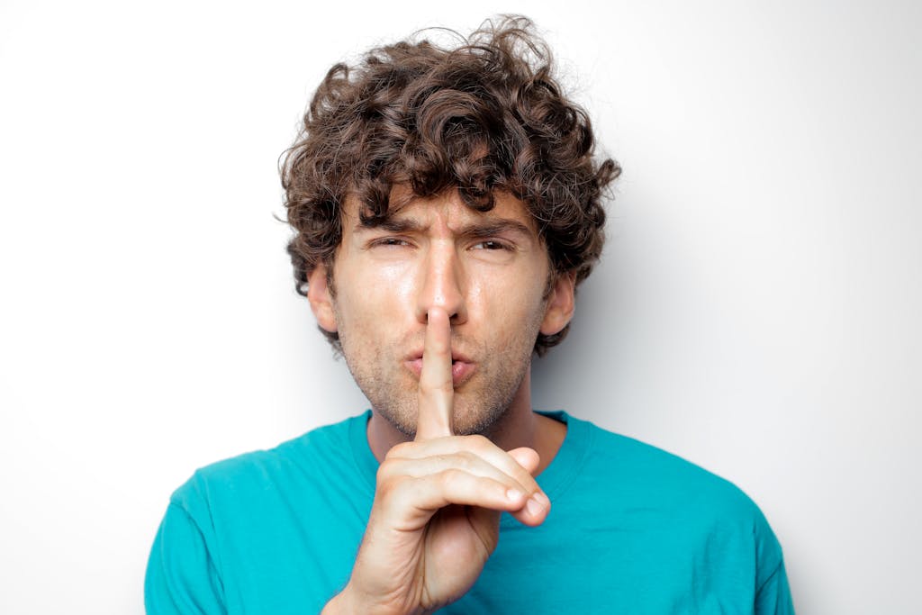 Adult man with curly hair in blue shirt making a silence gesture on white background.