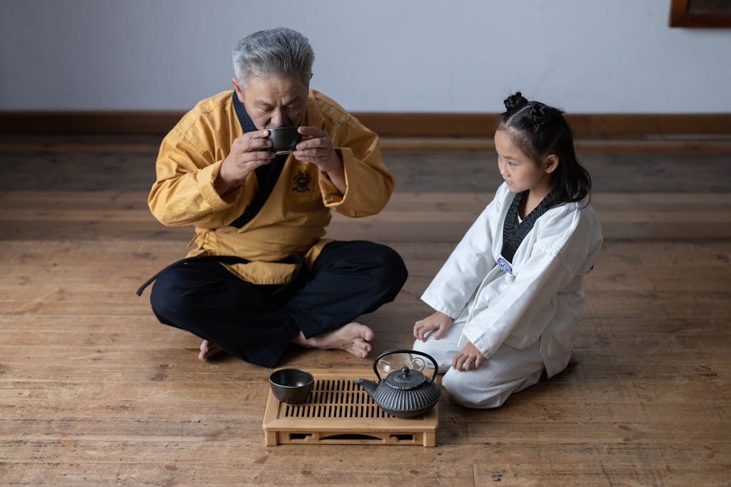 Elderly man and young girl in martial arts uniforms participate in a traditional tea ceremony.