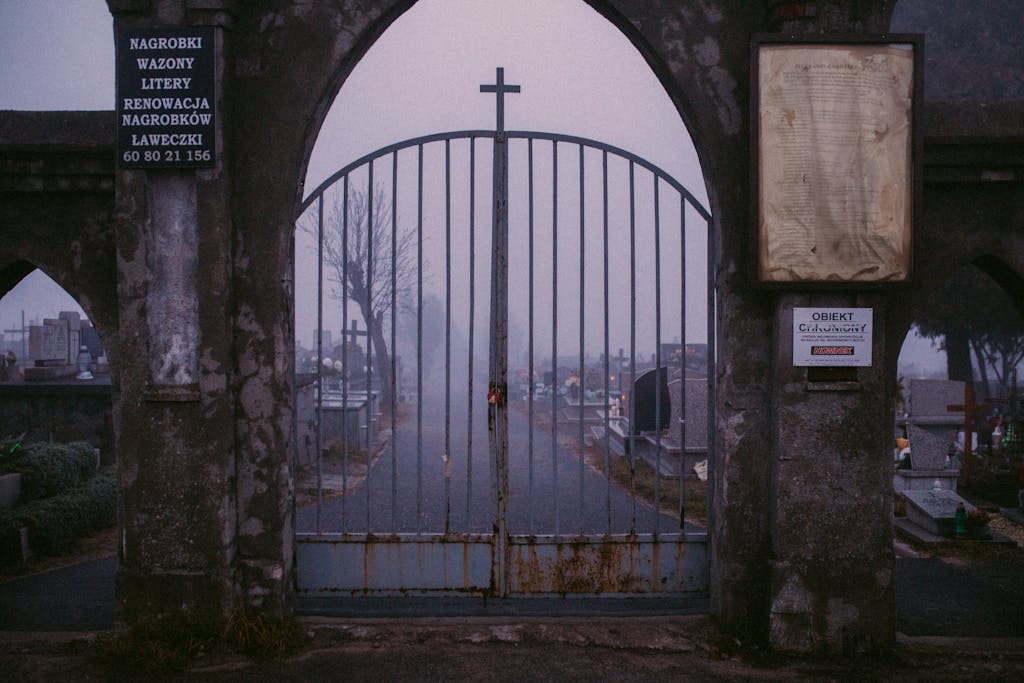 A mysterious foggy cemetery entrance with rusty iron gate and cross, creating an eerie atmosphere.