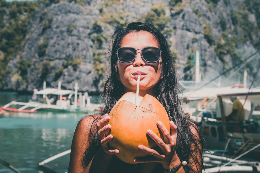 A woman in sunglasses enjoying a fresh coconut drink by the water, with boats and mountains in the background.