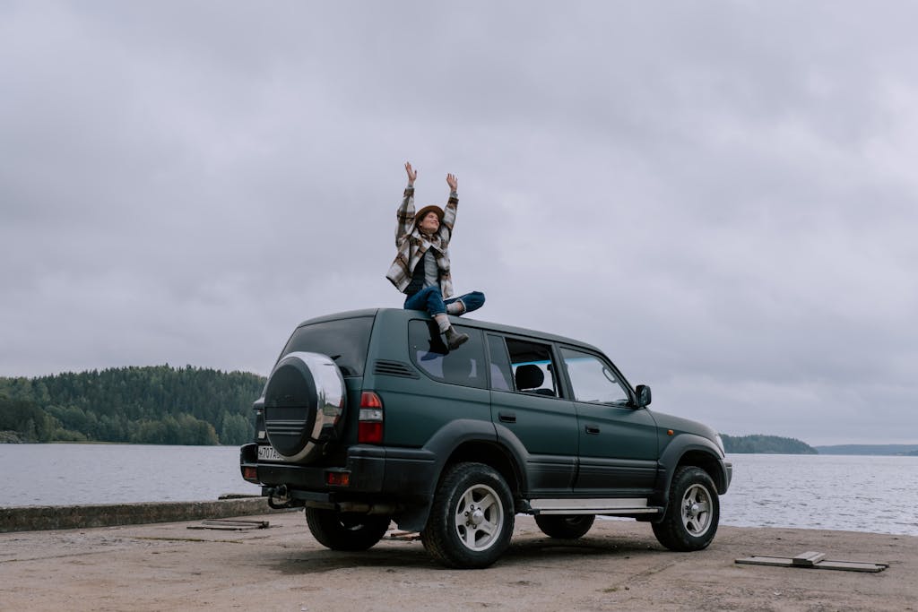 A woman sits on top of an SUV with arms raised in excitement by a scenic lake.