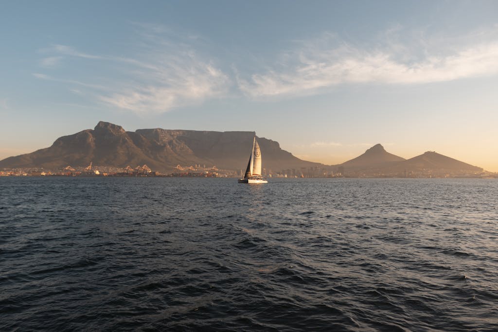 Sailboat navigating the ocean with Table Mountain and Cape Town in the backdrop during a serene sunset.